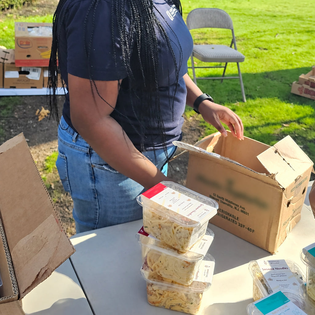 A woman with long black braids and a black headscarf stands at an outdoor table, packing food items into a cardboard box. The scene is set in a grassy park area with trees and buildings in the background under a blue sky.